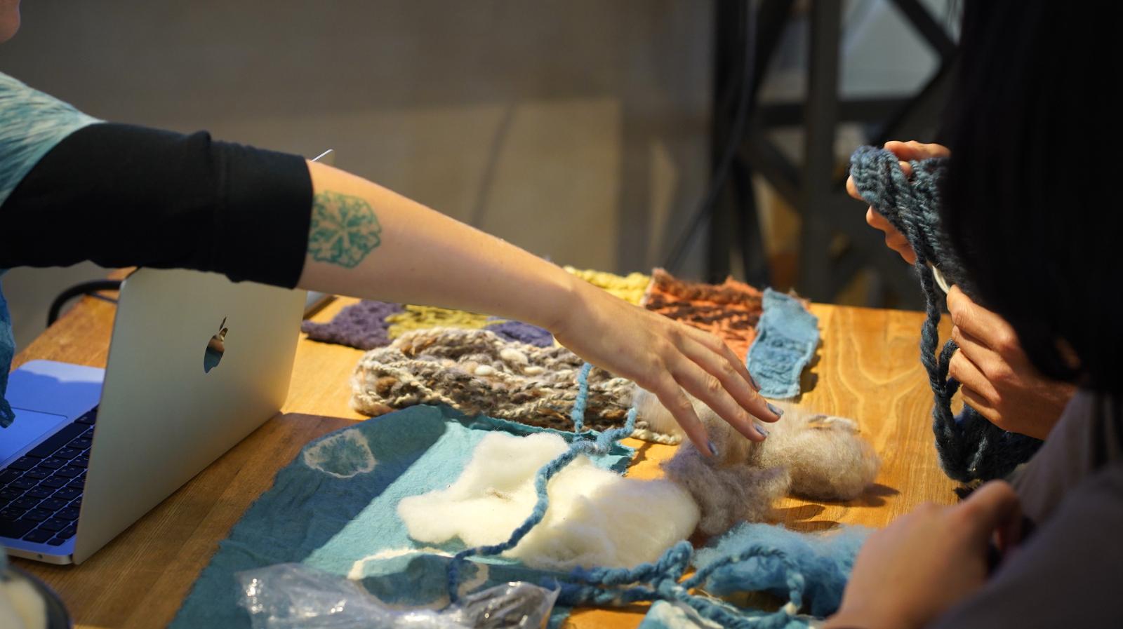 A photo of several hand-woven and dyed textiles. Three people gesture toward or hold samples.